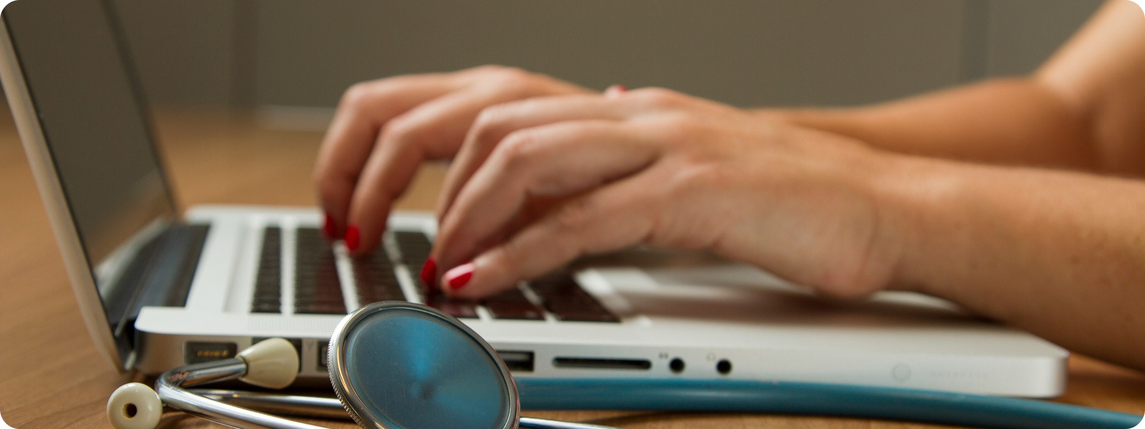 A doctor typing on a laptop, a stethoscope resting on the desk beside it